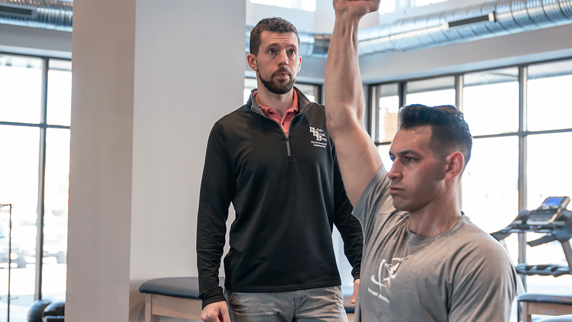 A man lifts a kettlebell over his head while his Physical Therapist observes.